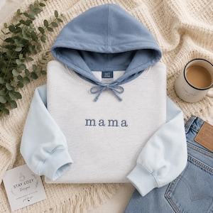 May include: A light gray hooded sweatshirt featuring a dusty blue hood and matching blue drawstrings. The word mama is embroidered in blue thread across the center of the chest. The sweatshirt has long sleeves with ribbed cuffs and a soft fabric texture.