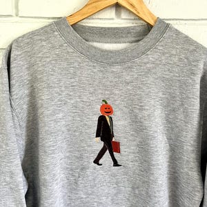 May include: A heather gray sweatshirt with an embroidered design of a person with a pumpkin head wearing a black suit and carrying a red briefcase. The design is centered on the chest.
