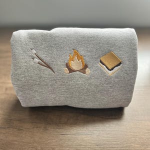 May include: A folded gray sweatshirt with embroidered camping-themed designs. The designs include a campfire, a s'more, and marshmallows on sticks. The sweatshirt is made of a soft material and is perfect for outdoor adventures.