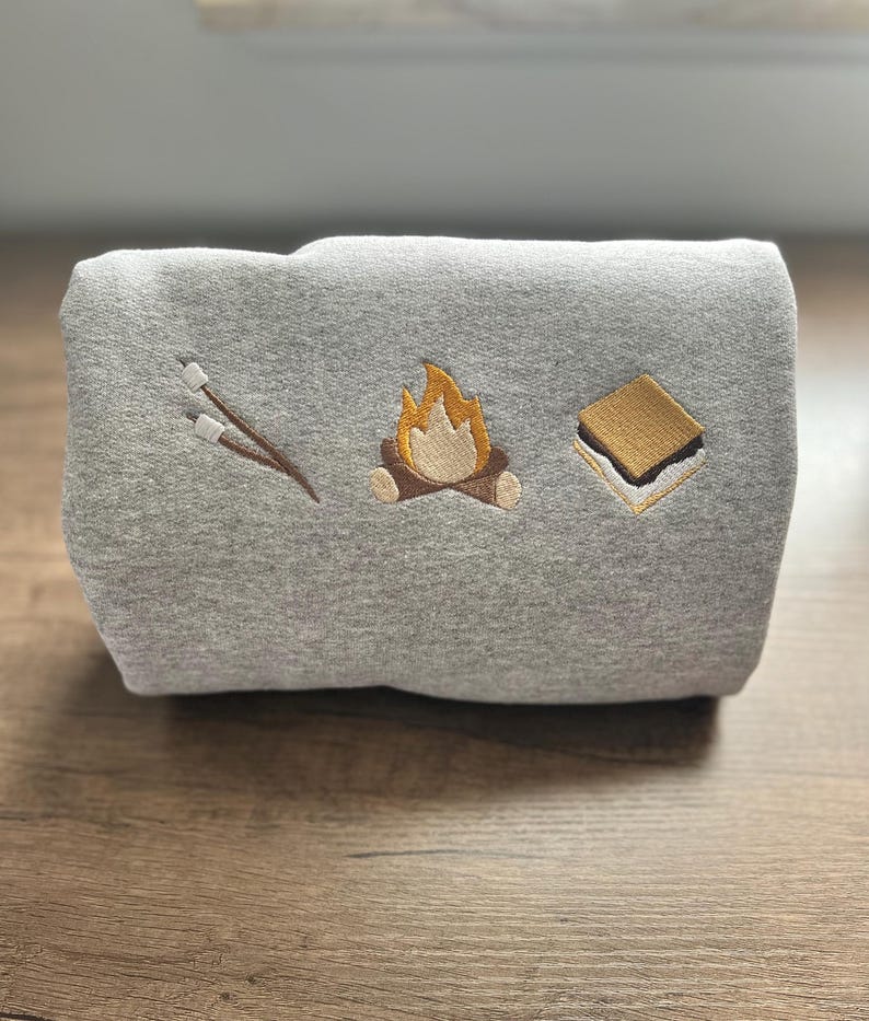 May include: A folded gray sweatshirt with embroidered camping-themed designs. The designs include a campfire, a s'more, and marshmallows on sticks. The sweatshirt is made of a soft material and is perfect for outdoor adventures.