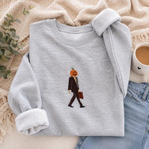 May include: A light gray crewneck sweatshirt featuring a small, centered embroidery of a figure with a pumpkin head wearing a dark brown suit and carrying a briefcase. The figure is depicted in a walking pose with a yellow tie and orange pumpkin.