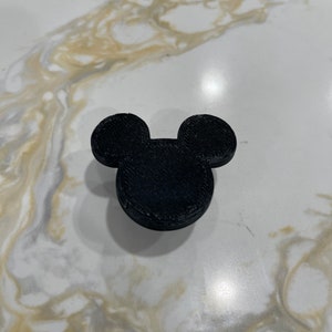 May include: A black 3D-printed Mickey Mouse head silhouette. The iconic character's ears and face are clearly defined. The object is sitting on a white and gold marble-patterned surface.