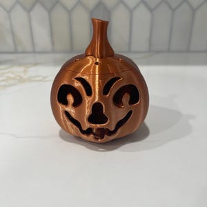 May include: A bronze-colored, 3D-printed pumpkin with a carved jack-o'-lantern face. The pumpkin has a stem and small holes on top. This decorative item is perfect for Halloween.
