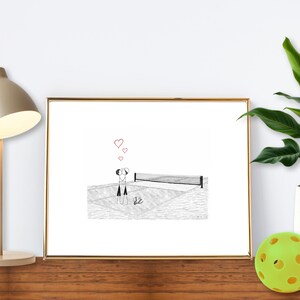 May include: A framed black and white illustration depicts two figures playing pickleball, with hearts above them. The artwork is displayed on a wooden surface, next to a lamp and a potted plant. Two yellow pickleballs are also present.