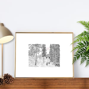 May include: A framed black and white illustration of two figures in a forest, with a heart above them. The artwork is in a gold frame, displayed on a wooden surface with a lamp and a fern in a pot.