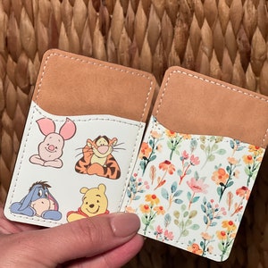 May include: Two brown leather card holders with white inserts. One card holder features a floral pattern with yellow, pink, and blue flowers. The other card holder features cartoon characters from Winnie the Pooh, including Piglet, Tigger, Eeyore, and Winnie the Pooh.