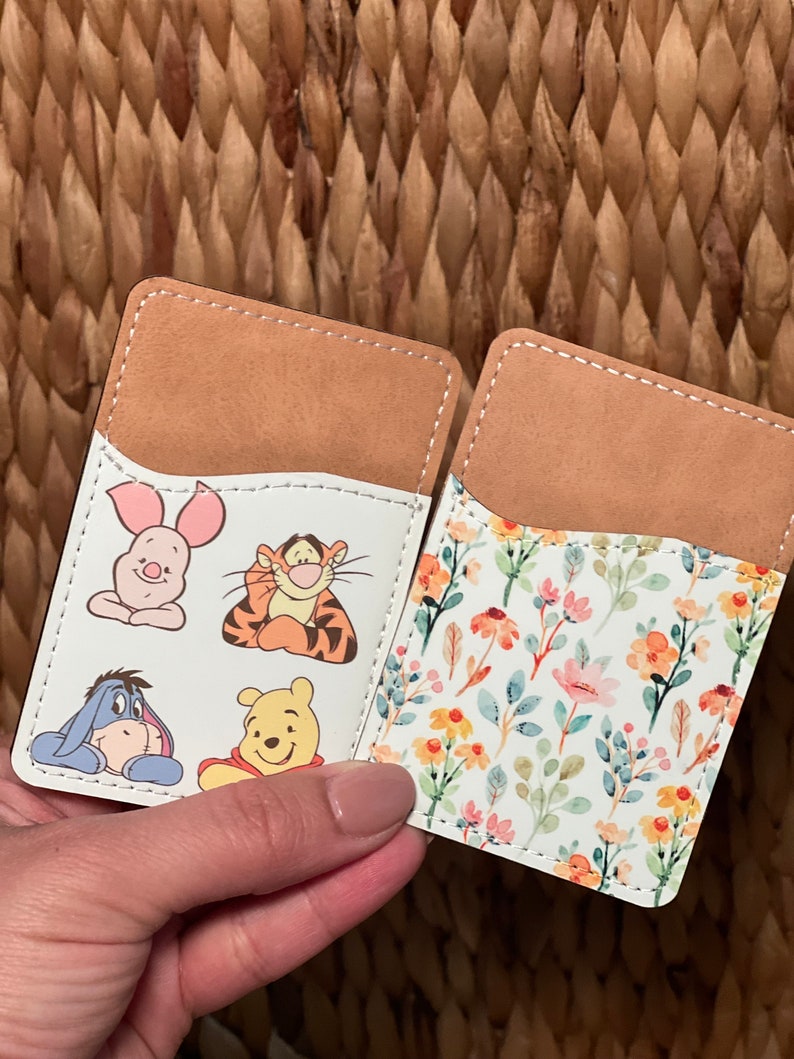 May include: Two brown leather card holders with white inserts. One card holder features a floral pattern with yellow, pink, and blue flowers. The other card holder features cartoon characters from Winnie the Pooh, including Piglet, Tigger, Eeyore, and Winnie the Pooh.