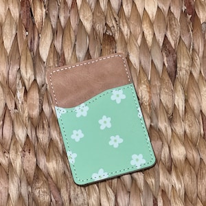 May include: A green leather card holder with a white floral pattern. The card holder has a brown leather flap on the back.