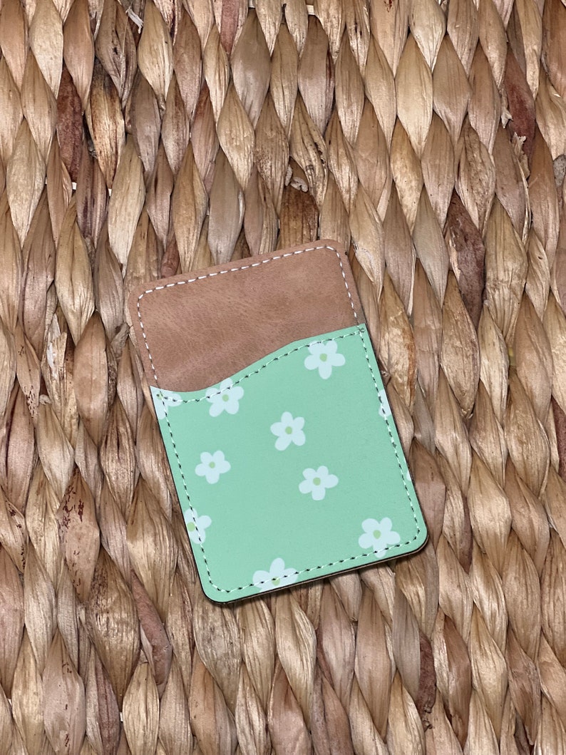 May include: A green leather card holder with a white floral pattern. The card holder has a brown leather flap on the back.