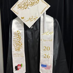 May include: A black graduation gown with a white cap and sash. The cap has gold calligraphy and a tassel. The sash features the text "Class of 2026" and "My Success is only by Allah", along with the flags of Afghanistan and the United States.