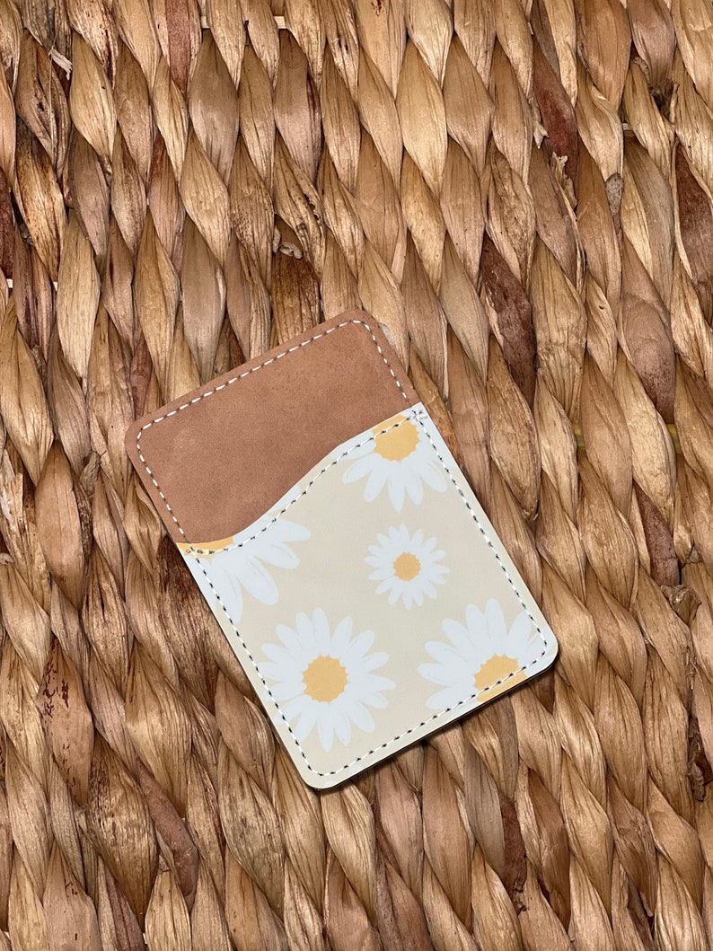 May include: A small brown leather wallet with a white and yellow daisy print. The wallet has a flap that folds over to close.