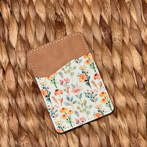 May include: A small, brown leather card holder with a white floral pattern. The pattern features pink, yellow, and green watercolor flowers and leaves.