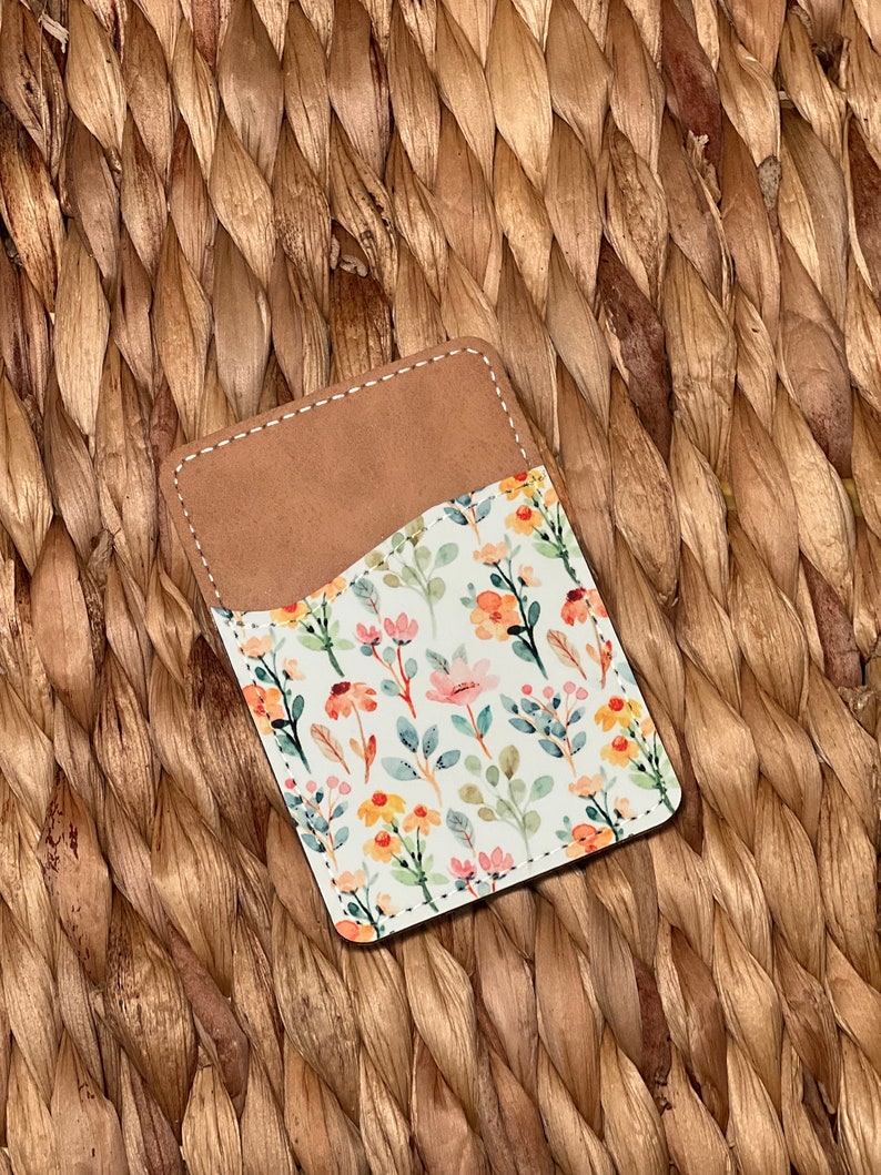 May include: A small, brown leather card holder with a white floral pattern. The pattern features pink, yellow, and green watercolor flowers and leaves.