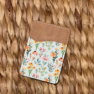 May include: A small, brown leather card holder with a white floral pattern. The pattern features pink, yellow, and green flowers and leaves.