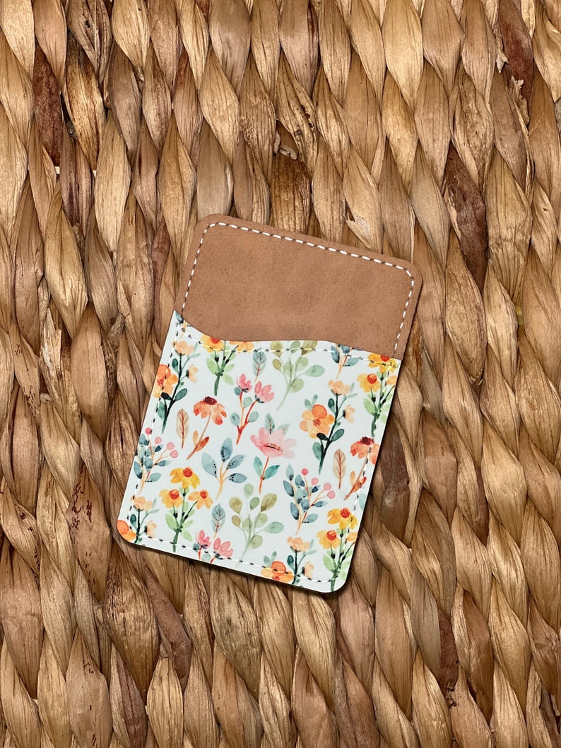 May include: A small, brown leather card holder with a white floral pattern. The pattern features pink, yellow, and green flowers and leaves.