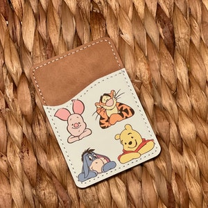 May include: A brown leather card holder with a white interior featuring cartoon images of Winnie the Pooh, Piglet, Tigger, and Eeyore.