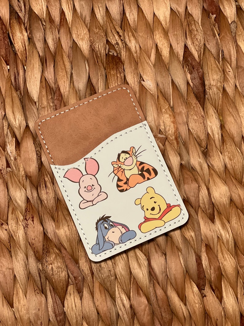 May include: A brown leather card holder with a white interior featuring cartoon images of Winnie the Pooh, Piglet, Tigger, and Eeyore.