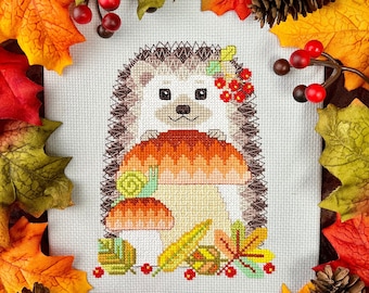 May include: A cross-stitch pattern of a brown hedgehog with a red and white mushroom on its back. The hedgehog is surrounded by green leaves and red berries. The pattern is on a white background.