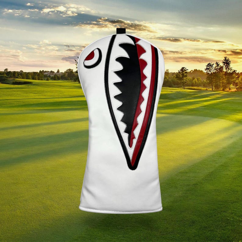 Op de afbeelding: Witte golfclubhoofdbeschermer met een haaienontwerp. De hoes heeft een haaienbek met zwarte en rode accenten, een rood oog en zwarte stiksels. De achtergrond toont een groene golfbaan onder een zonsondergang.