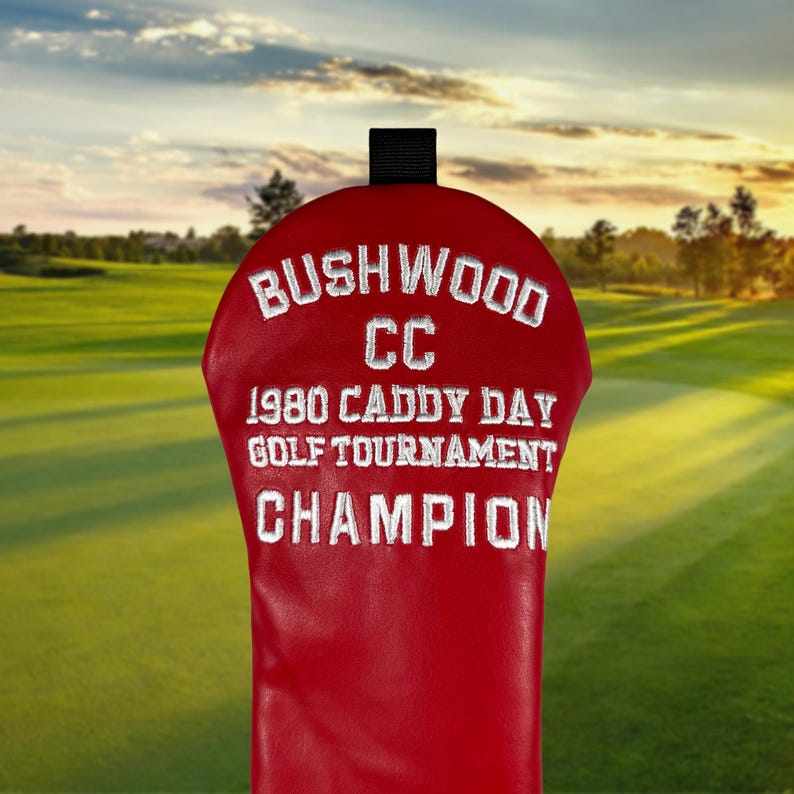 以下が含まれることがあります： 白い刺繍のテキストが入った赤いゴルフクラブヘッドカバー: "Bushwood CC 1980 Caddy Day Golf Tournament Champion." カバーの上部には黒いループが付いています。背景はゴルフコースです。