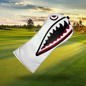 Op de afbeelding: Witte golfclubhoofdbeschermer met een haaienontwerp. De hoes heeft een zwart en rood open mondontwerp met scherpe tanden. De hoes staat tegen een groene golfbaanachtergrond.