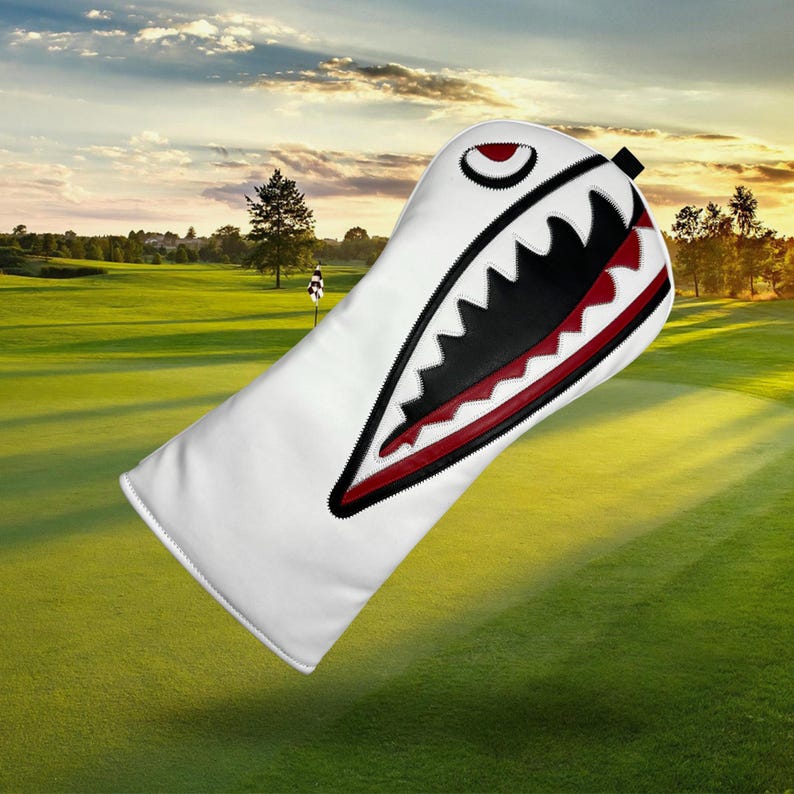 Op de afbeelding: Witte golfclubhoofdbeschermer met een haaienontwerp. De hoes heeft een zwart en rood open mondontwerp met scherpe tanden. De hoes staat tegen een groene golfbaanachtergrond.