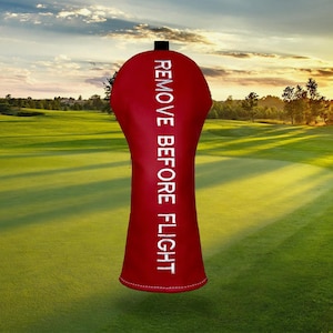 以下が含まれることがあります： 白い文字で「REMOVE BEFORE FLIGHT」と書かれた赤いゴルフクラブヘッドカバー。カバーは滑らかな素材でできており、黒いトップが付いています。背景は、緑の芝生と夕焼けのあるゴルフコースです。