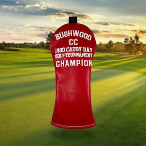 以下が含まれることがあります： 「Bushwood CC 1980 Caddy Day Golf Tournament Champion」と白い刺繍が施された赤いゴルフクラブヘッドカバー。カバーは黒いトップで、ゴルフコースの背景に設定されています。