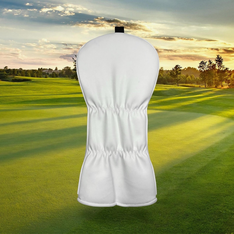 Op de afbeelding: Een witte golfclubhoofdbeschermer met een zwarte bovenkant. De hoes is gemaakt van een glad materiaal en heeft een gestikt ontwerp. De achtergrond is een golfbaan met groen gras en een zonsondergang.
