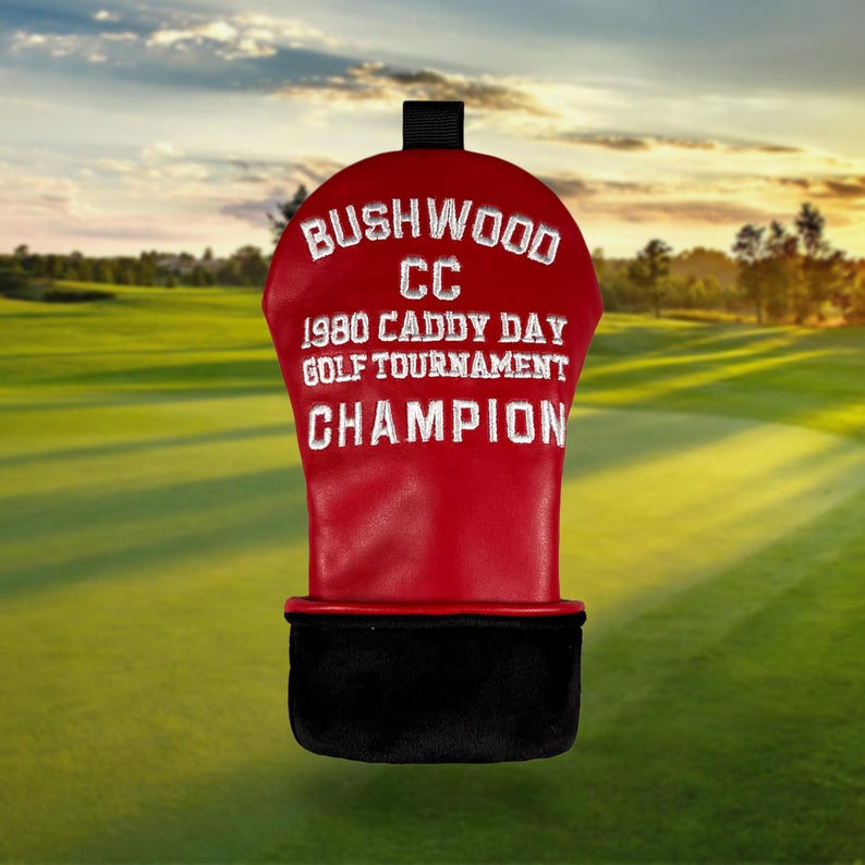 以下が含まれることがあります： 「Bushwood CC 1980 Caddy Day Golf Tournament Champion」と白い刺繍が施された赤いゴルフクラブヘッドカバー。底は黒で、上部に黒いループが付いています。背景はゴルフコースです。