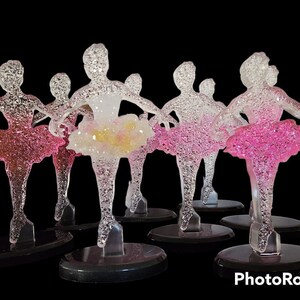 Könnte beinhalten: Eine Gruppe von sieben klaren Glas-Ballerina-Figuren mit rosa und weißen Glitzer-Tutus. Die Figuren stehen auf schwarzen Sockeln.
