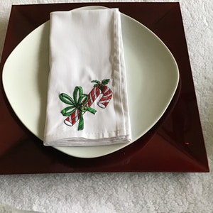 May include: White linen napkin with a red and white candy cane and green bow embroidery design. The napkin is folded and placed on a white plate with a red charger.