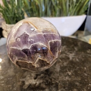 May include: A clear crystal ball with a mosaic of purple amethyst pieces. The sphere has a polished surface and sits on a dark, textured surface. The background includes a white bowl and green and purple plants.