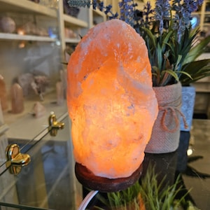 May include: A glowing orange Himalayan salt lamp on a dark wooden base. The lamp is illuminated from within, casting a warm light. A white power cord extends from the base. The background includes shelves with crystals and a vase of lavender.