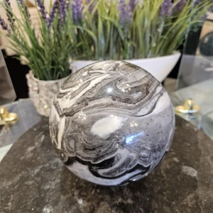 May include: A polished stone sphere with a swirling pattern of black, white, and gray. The sphere is displayed on a dark, textured surface. Decorative lavender plants in white pots are in the background.