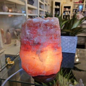 May include: A glowing Himalayan salt lamp. The lamp is a large, rough-hewn chunk of pink and orange salt, emitting a warm, inviting light. It sits on a dark wooden base and is plugged in with a white cord. The lamp is a decorative and functional item.