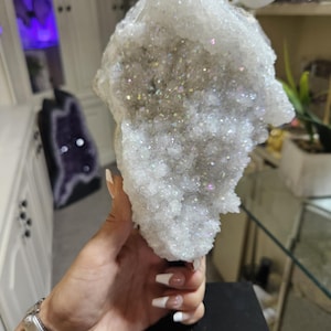 May include: A large, iridescent quartz crystal cluster. The crystal is a light, translucent color with sparkling, rainbow-like inclusions. The cluster is held against a blurred background, showcasing its natural, textured surface. The crystal is a unique, decorative item.