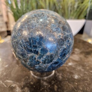 May include: A blue apatite sphere with a cracked, textured surface. The gemstone ball is displayed on a clear stand, showcasing its intricate patterns and color variations. The sphere is a decorative item.