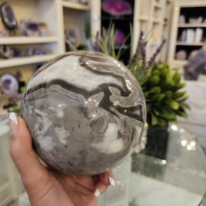 May include: A polished sphere of banded agate, featuring swirling patterns of gray, white, and black. The gemstone is held in a hand, showcasing its smooth, rounded form. The sphere is a decorative item, likely for display or collection.