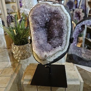 May include: A large, upright amethyst geode featuring deep purple, crystalline structures within a dark, rocky exterior. The geode is securely mounted on a black metal stand with a square base, displaying its natural, jagged, and sparkling interior.
