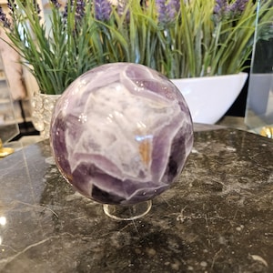 May include: A polished amethyst crystal sphere, showcasing a blend of purple and white hues. The sphere is displayed on a clear acrylic stand, set against a dark, marbled surface. Decorative lavender plants are visible in the background.