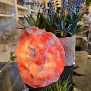 May include: A glowing, orange Himalayan salt lamp on a dark brown base. The lamp is next to a small potted plant with purple flowers and green leaves. The lamp is on a glass surface.
