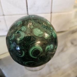 May include: A green malachite sphere with a swirling pattern. The sphere is sitting on a clear glass stand.