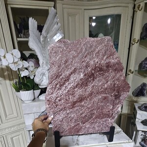 May include: A large, flat, irregular slab of lepidolite mineral with a shimmering, crystalline surface in shades of dusty rose and mauve. The stone is mounted on a simple black metal stand, displaying its natural, rough-hewn edges and texture.