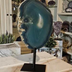 May include: A polished, translucent teal agate slice with concentric banding and a rough, textured outer edge. The stone is mounted vertically on a thin, black metal rod attached to a rectangular black metal base for display purposes.