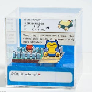 May include: A clear acrylic cube featuring a pixelated Snorlax figure from the Pokémon series. The figure is yellow and blue, standing on a wooden dock. The cube includes text that reads "SLEEPING POKÉMON" and "SNORLAX woke up!"