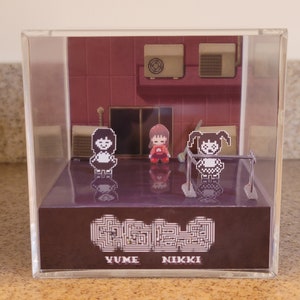 Può includere: Una teca in acrilico trasparente con un diorama in miniatura di una scena di videogioco. La scena presenta tre personaggi pixelati, un edificio rosso con condizionatori d'aria e il testo "Yume Nikki".