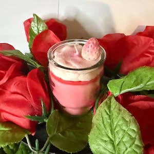 May include: A pink layered candle in a glass jar, with a strawberry wax topper. The candle is set amongst red roses and green leaves, creating a decorative and romantic display.