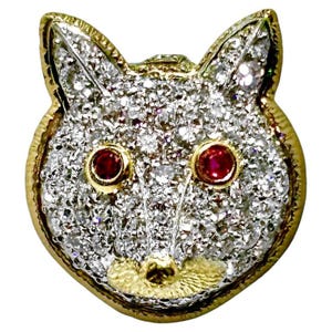 May include: A fox-shaped brooch crafted from gold and silver-toned metal, featuring a dense pavé setting of clear, sparkling stones across the face. The animal's eyes are set with two round, vibrant red stones, and the nose area has a textured gold finish.