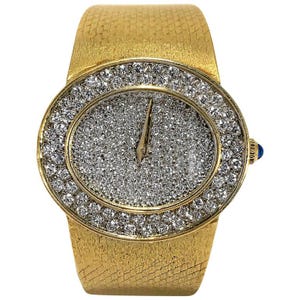 May include: A gold-toned wristwatch with a wide band and an oval face. The watch face is covered in small, sparkling stones, surrounded by a ring of larger stones. The watch has a small blue accent on the side.